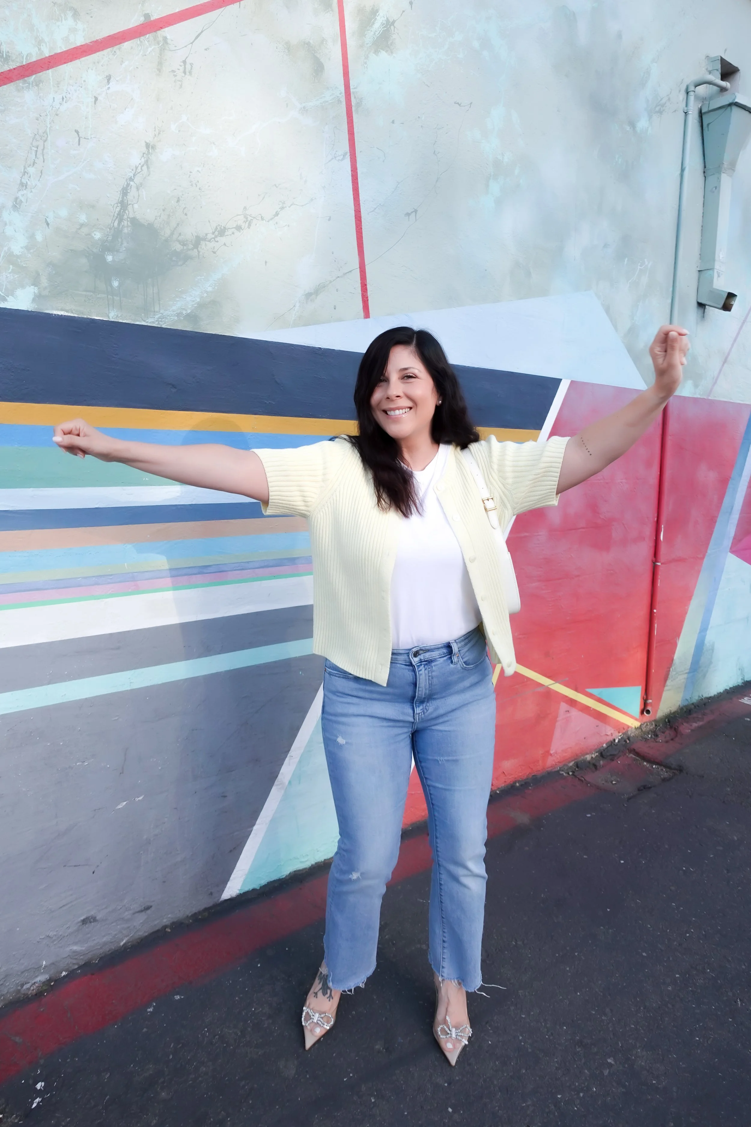 A woman poses smiling in front of a vibrant geometric street art mural. She wears a short-sleeve pale yellow cardigan over a white t-shirt, light-wash blue jeans, and clear heels with bow details. She is carrying a small white shoulder bag.