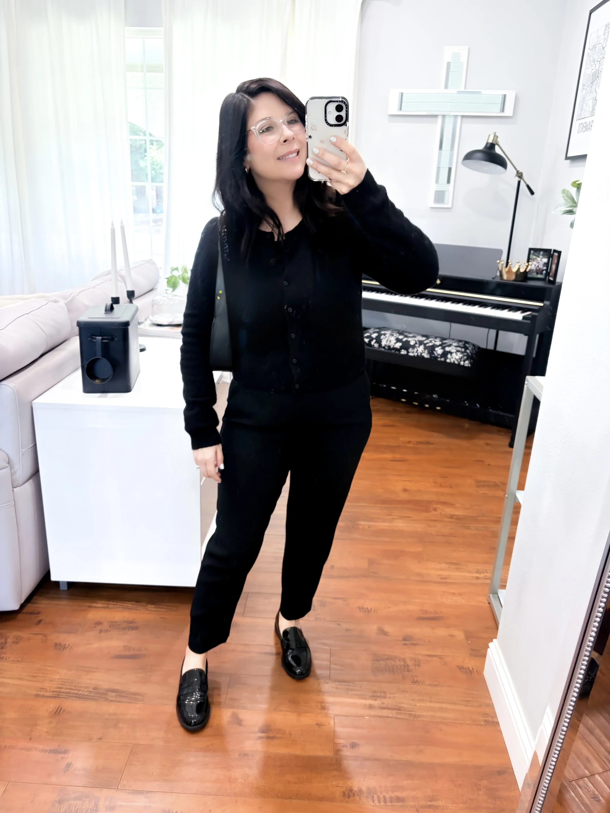 A classic monochromatic look featuring a black button-up cardigan, black ankle-length pants, and glossy black loafers, set in a brightly lit room with a piano.