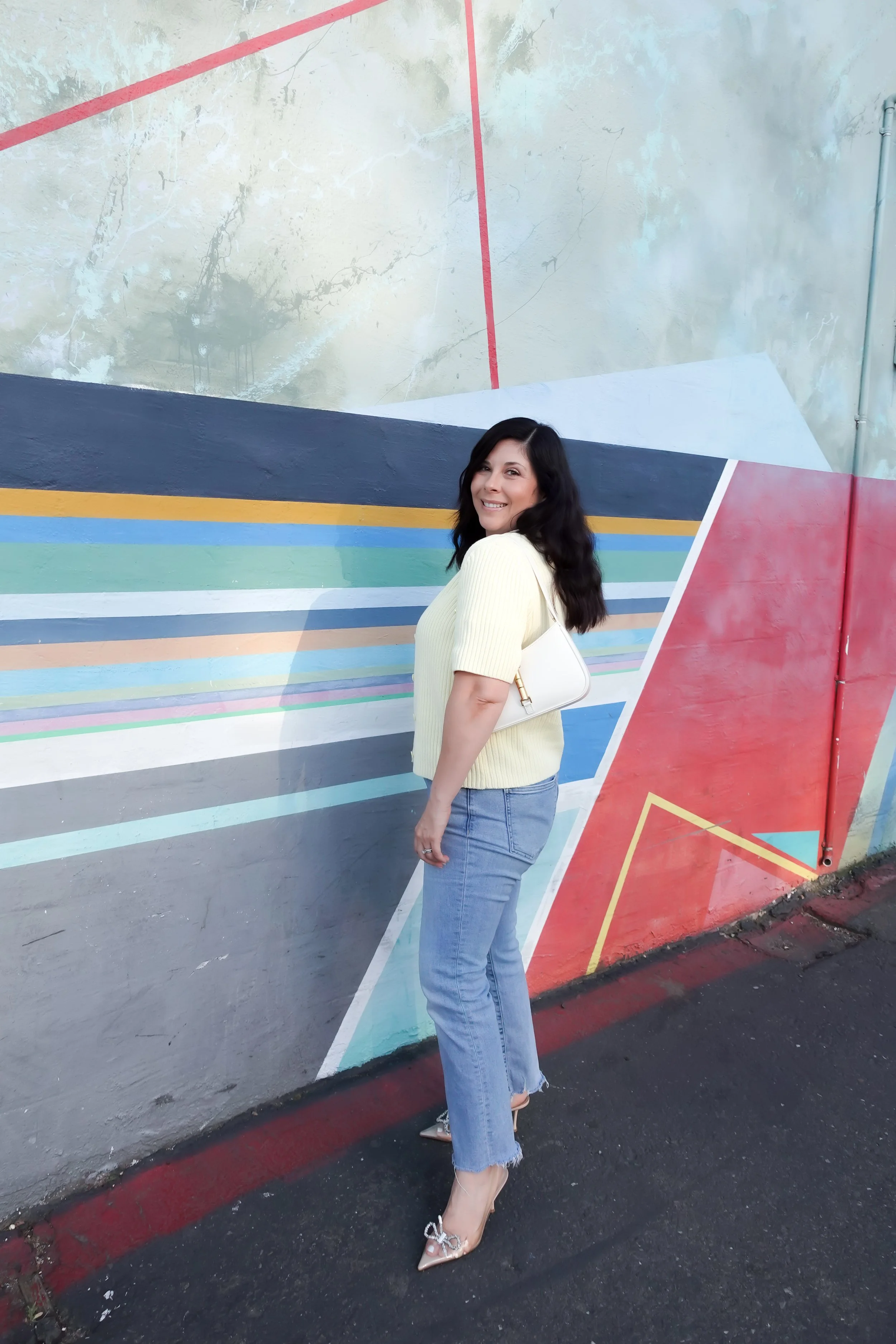 A woman poses smiling in front of a vibrant geometric street art mural. She wears a short-sleeve pale yellow cardigan over a white t-shirt, light-wash blue jeans, and clear heels with bow details. She is carrying a small white shoulder bag.