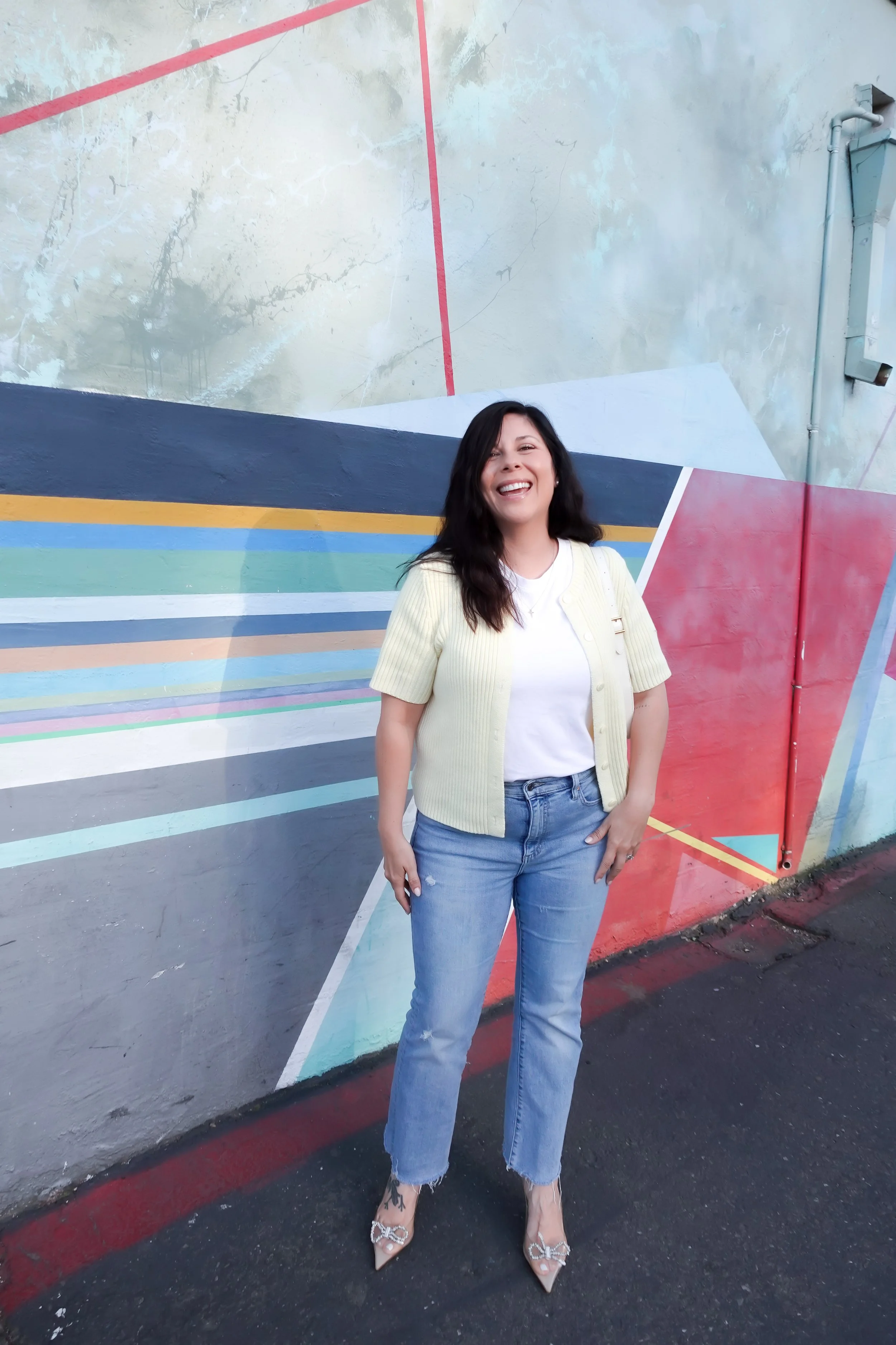 A woman poses smiling in front of a vibrant geometric street art mural. She wears a short-sleeve pale yellow cardigan over a white t-shirt, light-wash blue jeans, and clear heels with bow details. She is carrying a small white shoulder bag.