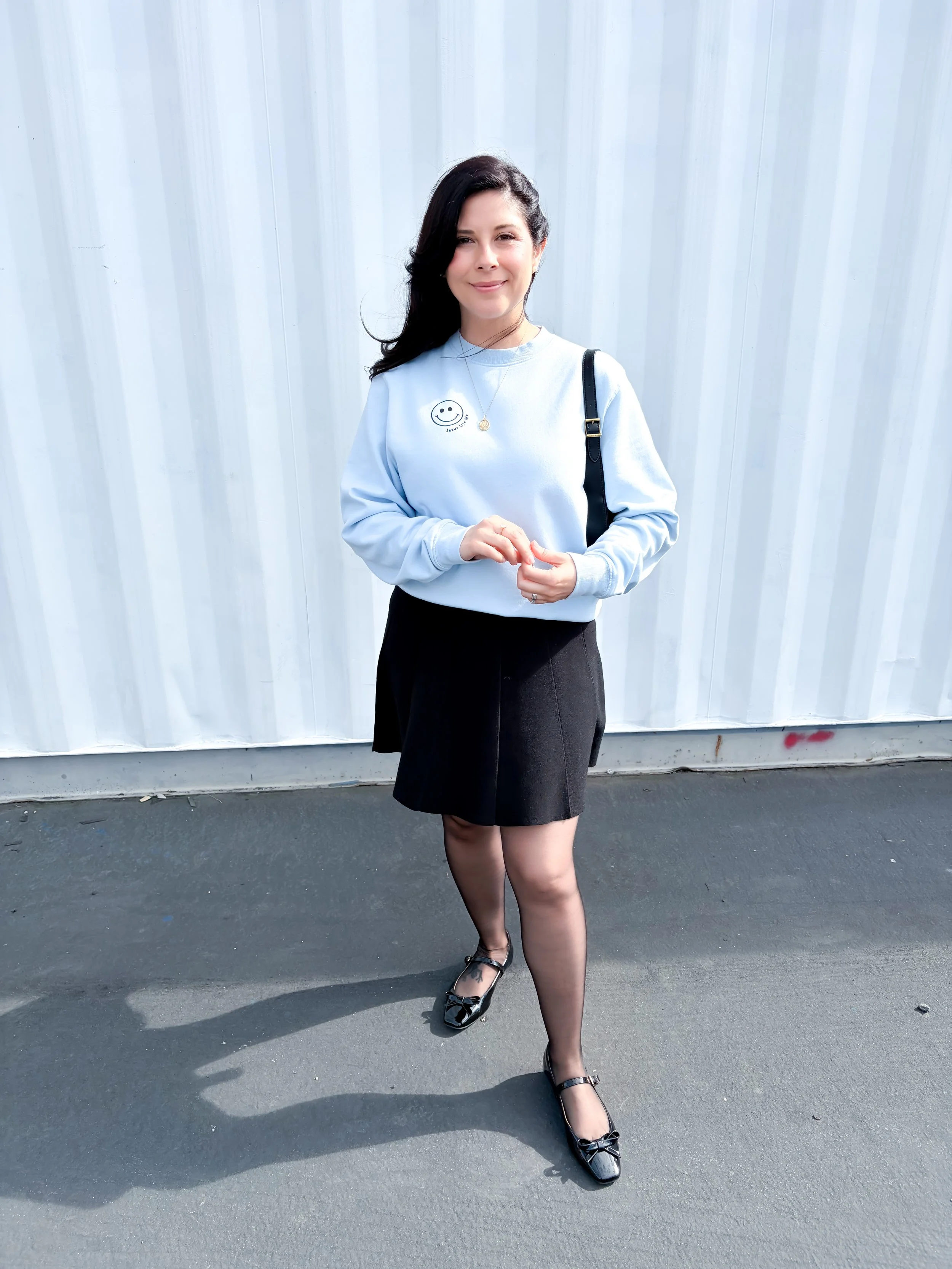 A classic spring outfit featuring a pastel blue crewneck sweatshirt, black skirt, and patent leather Mary Jane shoes.