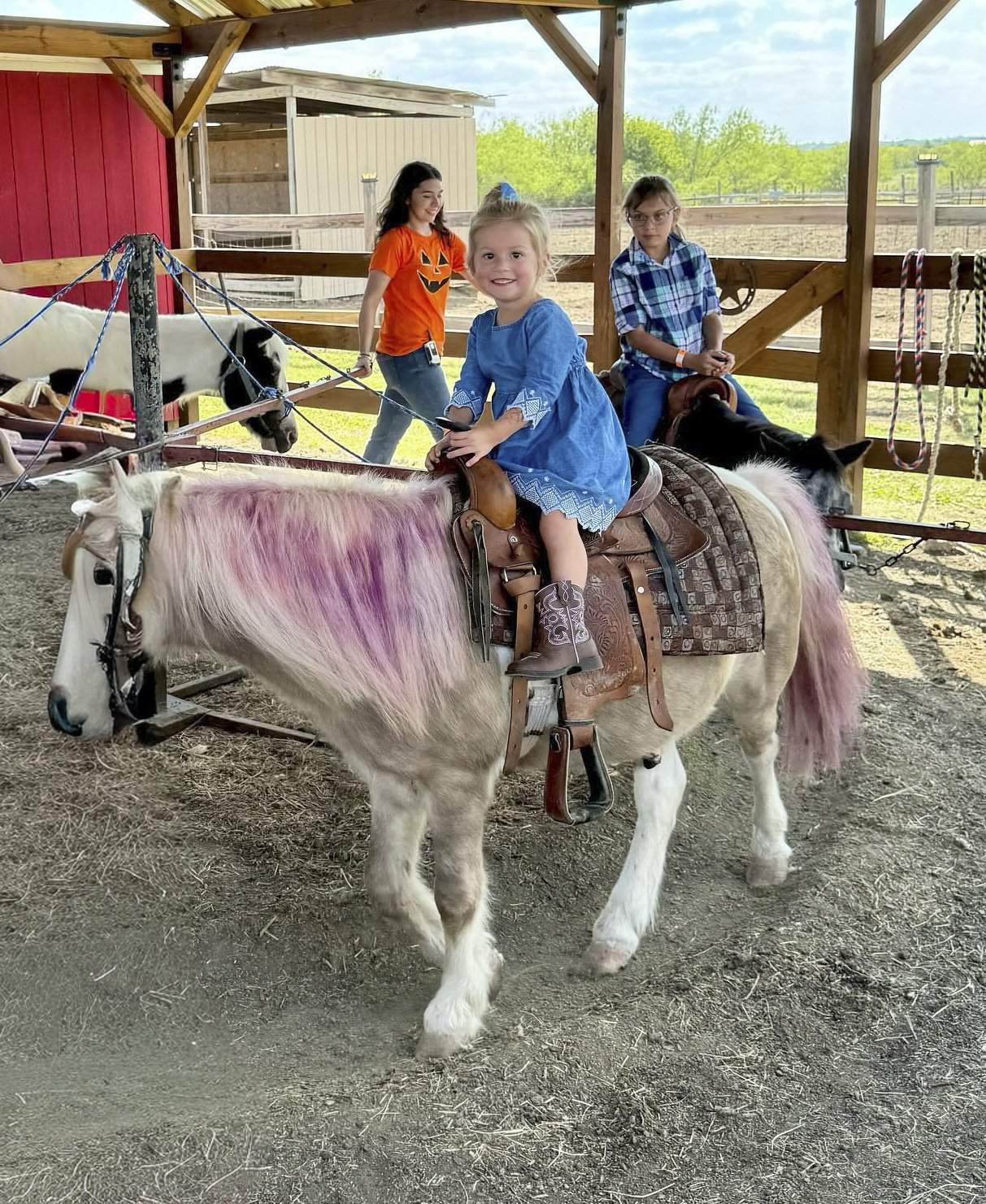 A young girl with blonde hair smiling while riding a light-colored horse in a paddock with a white picket fence.