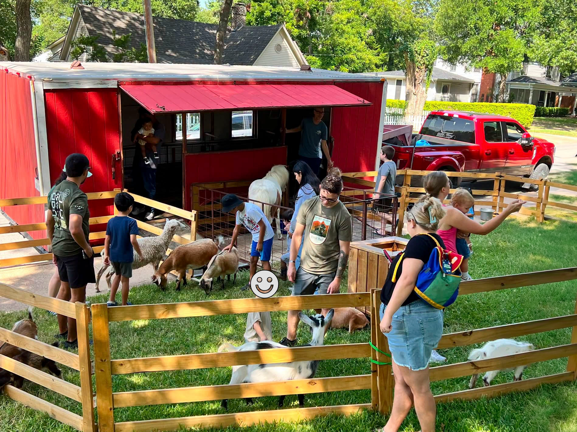 A small farm or petting zoo scene with a fenced area containing goats and a large red barn or trailer in the background.