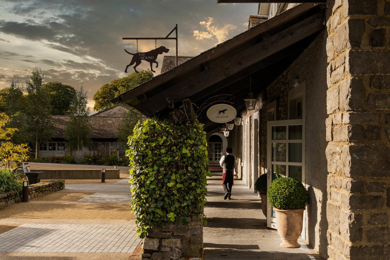 The exterior of a building with stone and brick walls, potted plants, and a hanging sign with a dog silhouette labeled "The Hound." A person dressed in black and white is walking along the sidewalk at sunset, with the sky partly cloudy and trees in t