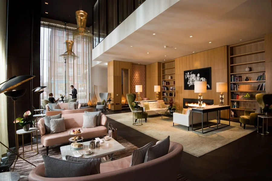 Modern hotel lobby with plush seating, large windows with curtains, and a fireplace. There are people sitting and conversing, with a reception desk in the background.
