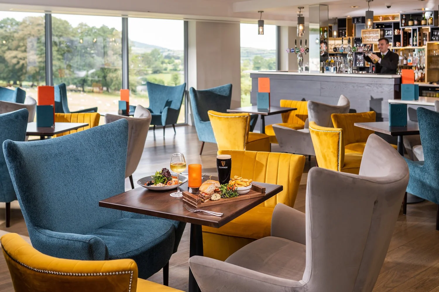 Interior of a colorful restaurant with chairs in yellow, blue, and beige, a table set with food and drinks, large windows with a view of a green landscape, and a bar area with a bartender.