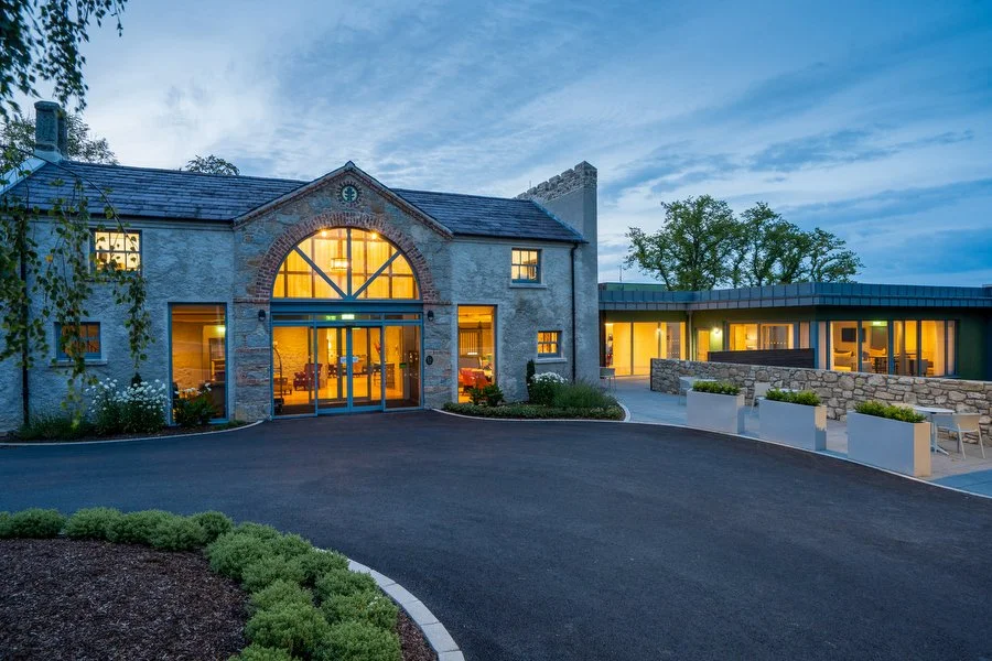 A well-lit building, Killeavy Castle, with an arched glass entrance and surrounding greenery during dusk.