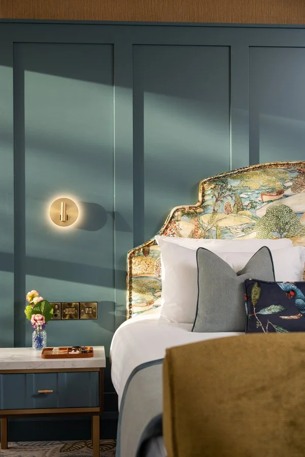 A bedroom corner featuring a bed with a decorative floral headboard, white linens, and gray accent pillows. Next to the bed is a blue bedside table with a small vase of pink and white flowers. The wall behind is painted blue with a modern wall sconce