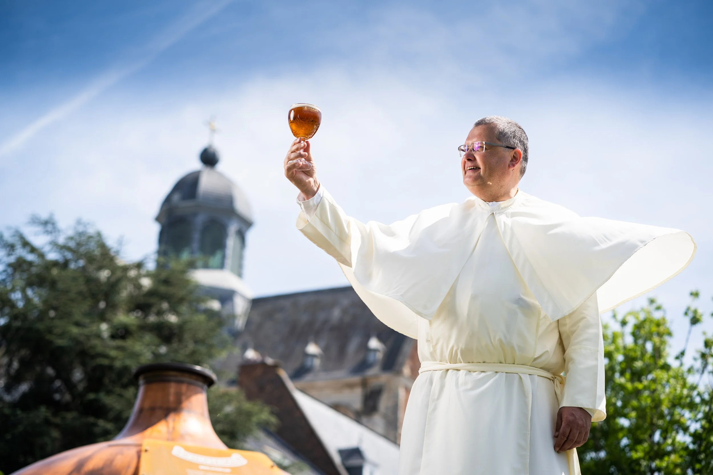 A priest wearing white robes holding up a glass of beer outdoors, with a church steeple and blue sky in the background.