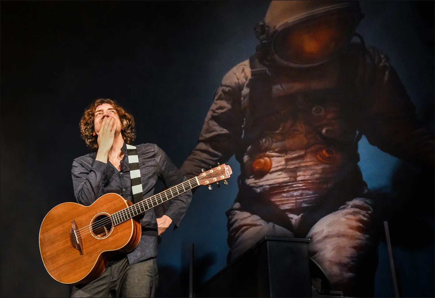A man with curly brown hair, dressed in a dark shirt, stands holding an acoustic guitar, covering his mouth with his hand, and appears to be in a museum or gallery, with a large astronaut painting or backdrop behind him.