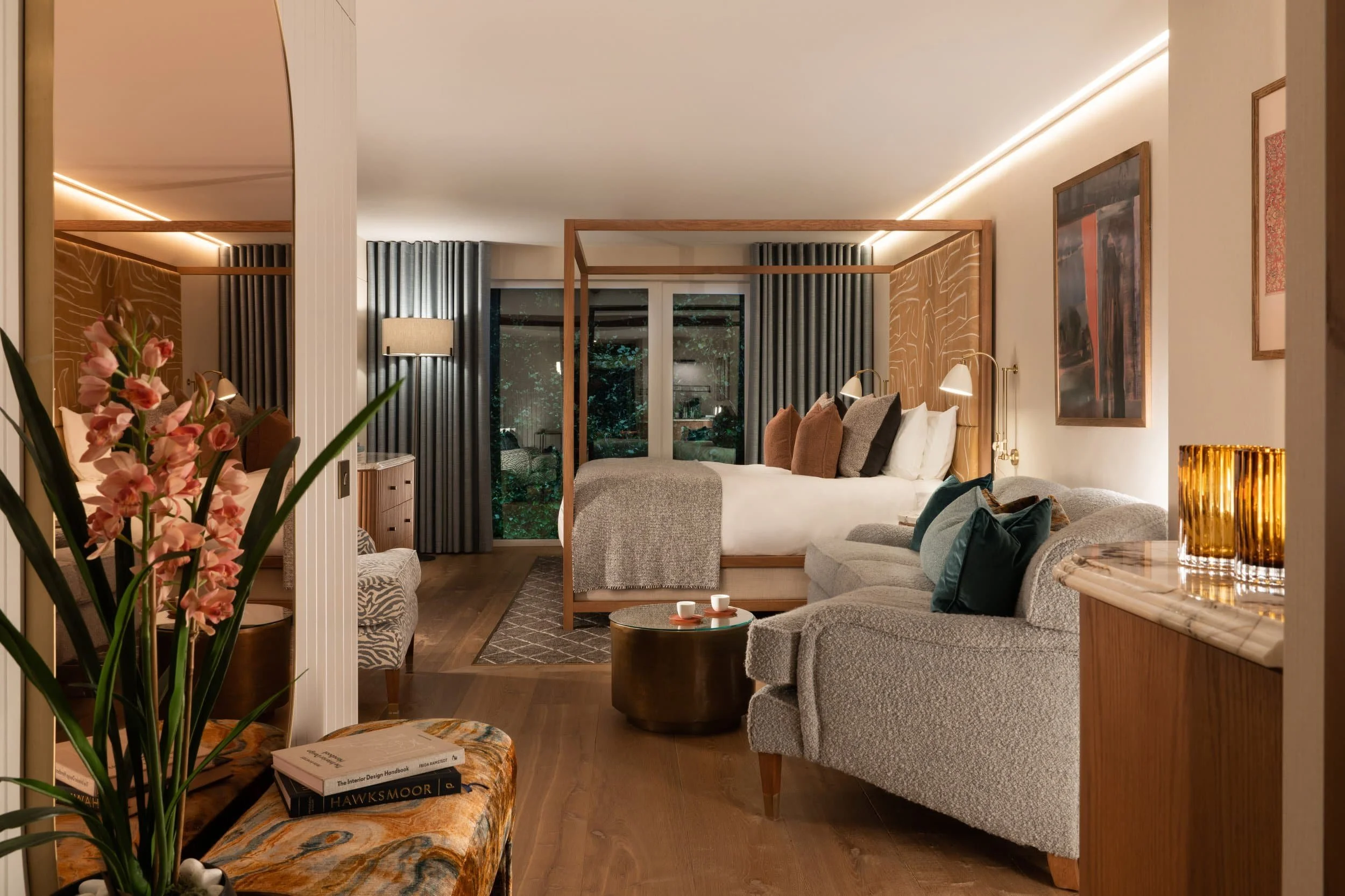 A luxurious hotel bedroom at the Clandeboye Lodge in Bangor, Co, Down with a four-poster bed, a seating area with a grey couch, a wooden coffee table with cups, and artwork on the walls, illuminated by warm lighting.