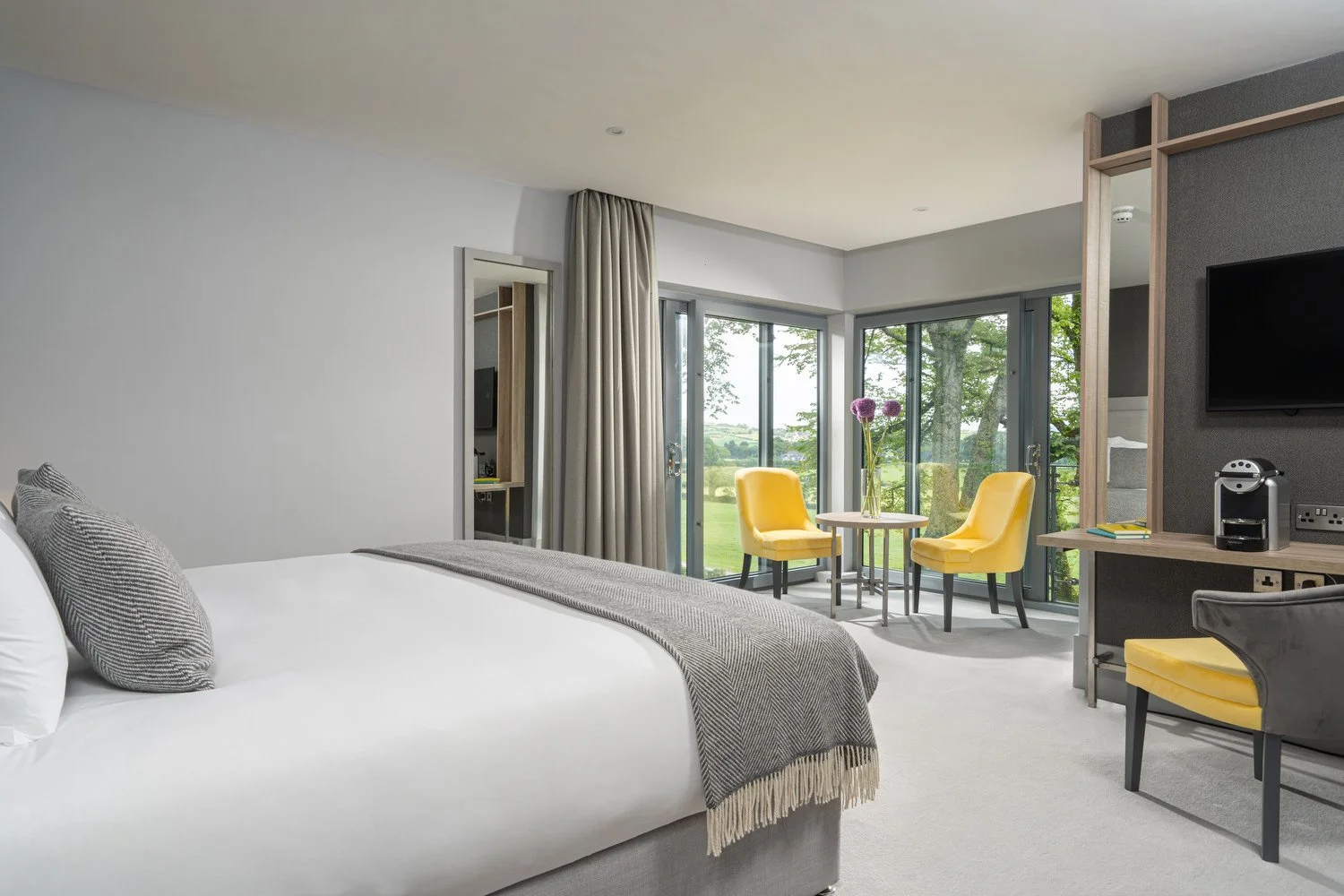 A modern hotel room with a large bed, gray and white color scheme, and a sitting area with two yellow chairs around a small round table near large sliding glass doors opening to a lush green outdoor view.