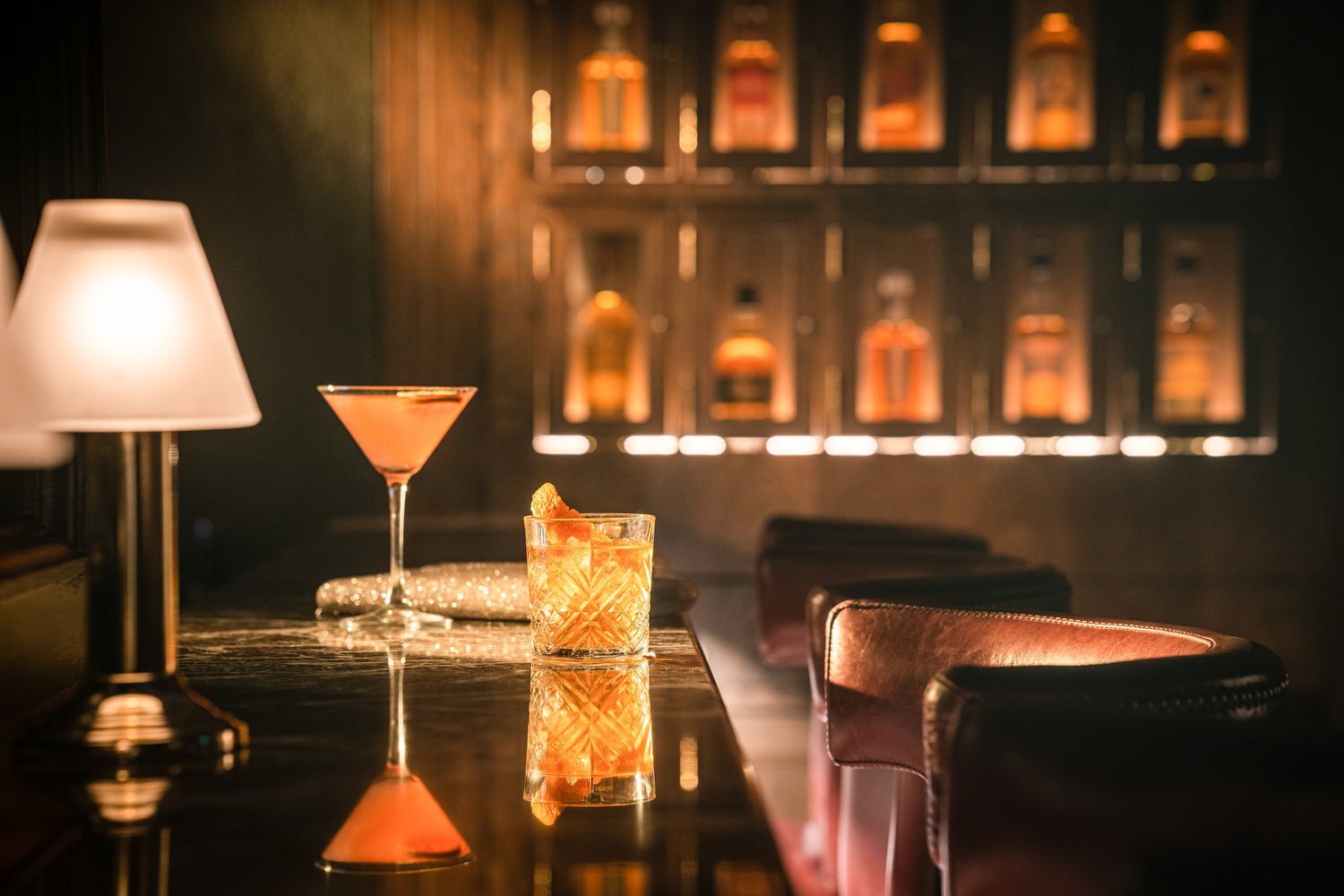 Inside a dimly lit bar with a wooden counter, vintage lamps, and three cocktails; one pink martini and one with an orange slice, reflecting on the glossy surface with leather chairs nearby and bottles displayed on illuminated shelves in the backgroun
