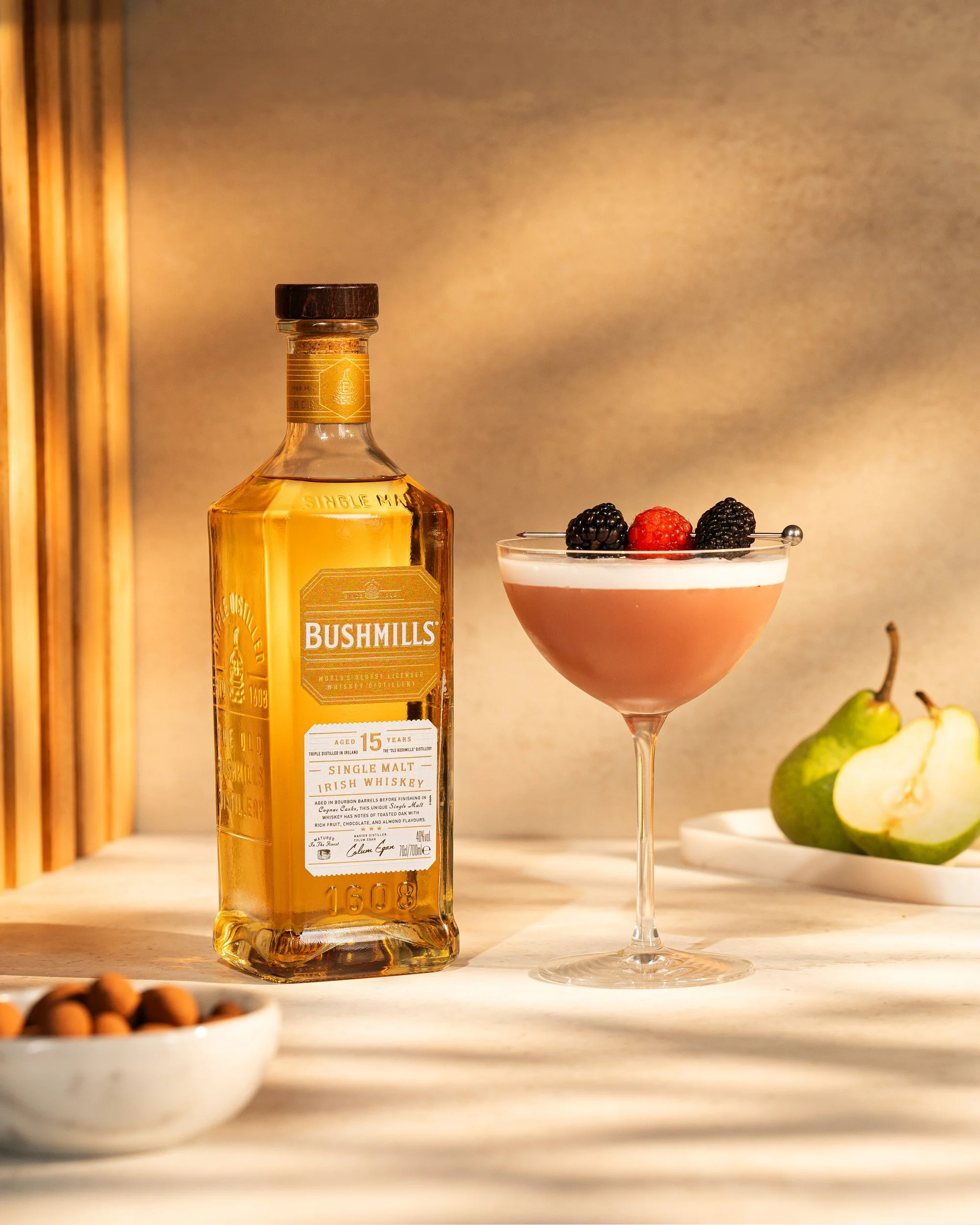 A bottle of Bushmills Irish whiskey, a pink cocktail garnished with blackberries and a raspberry, and a sliced pear on a white plate, with a bowl of nuts in the foreground.