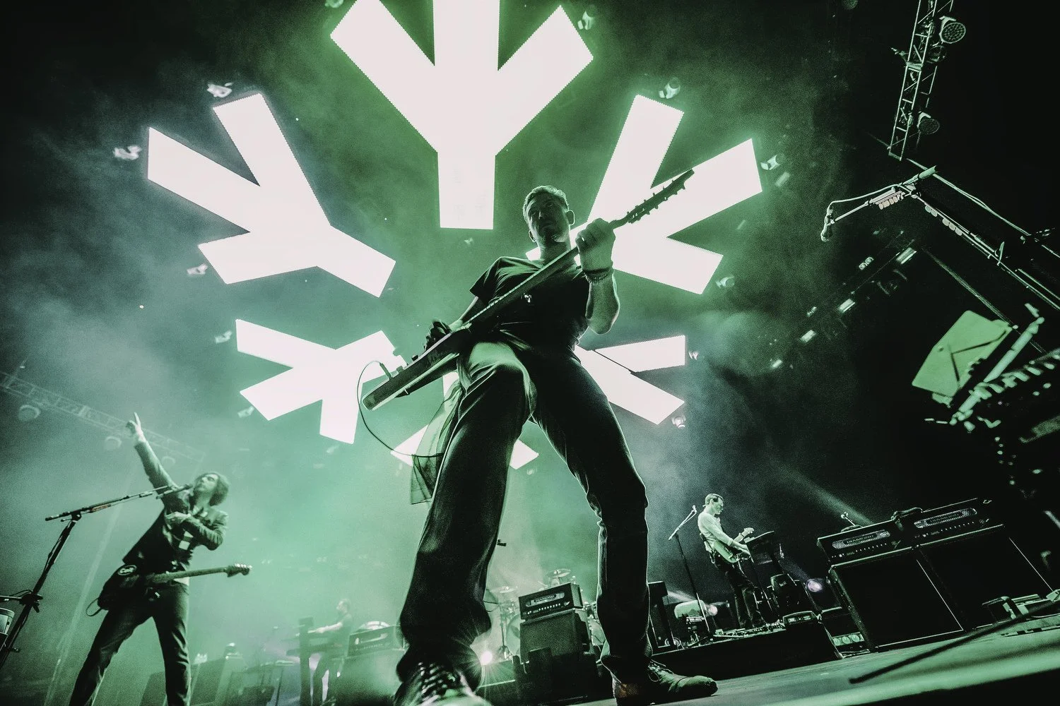 A band performs on stage with bright green lighting and an abstract light display in the background. The main guitarist at the center is holding an electric guitar, and other band members are playing instruments, with one member on the left pointing 