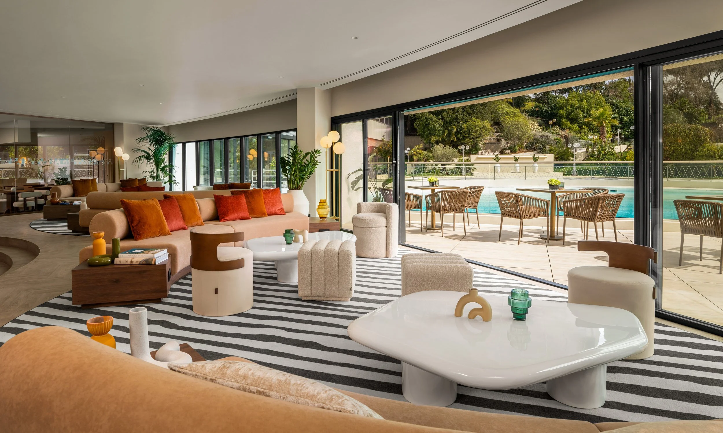 Modern hotel lobby with large glass doors opening to an outdoor pool area, featuring beige and brown furniture, striped black-and-white rug, and lush greenery.