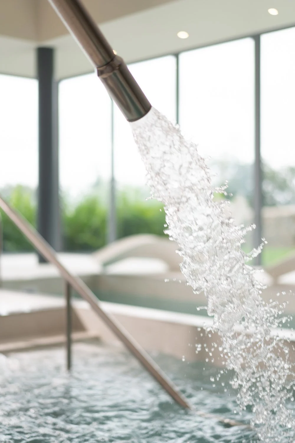 A spout of water flowing into a hot tub or pool in a bright indoor setting with large windows and a view of greenery outside.