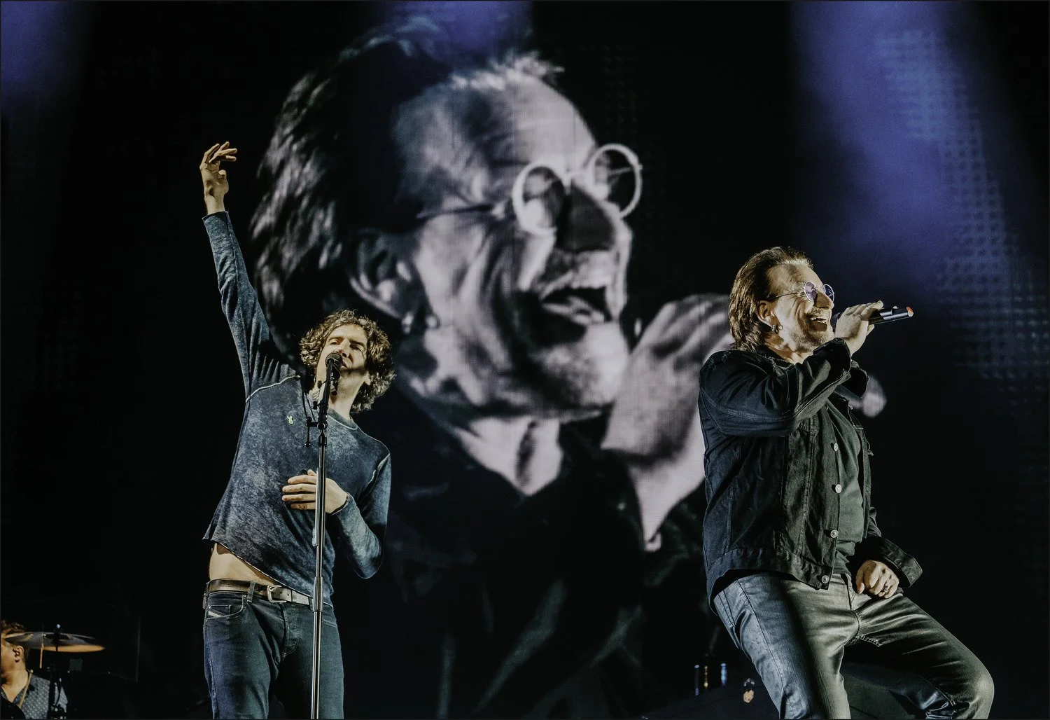 Two men singing on stage, one with curly hair and wearing a long sleeve shirt, the other with glasses and a jacket, both holding microphones, with a large black and white image of a woman singing projected behind them.