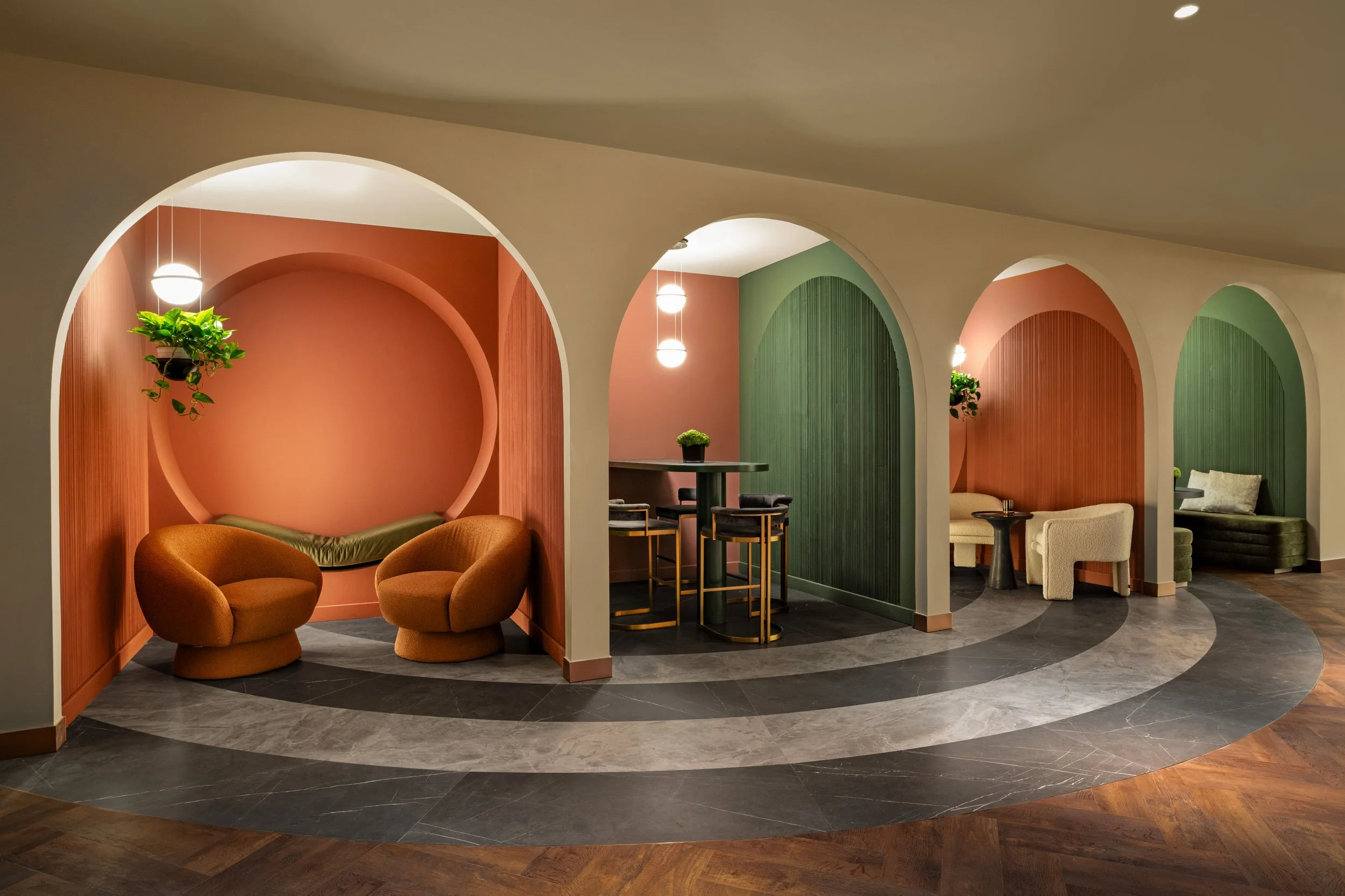 Modern lounge area with pink and green wall niches, featuring curved seating and hanging plants, illuminated by pendant lights, with a patterned marble and wood floor.