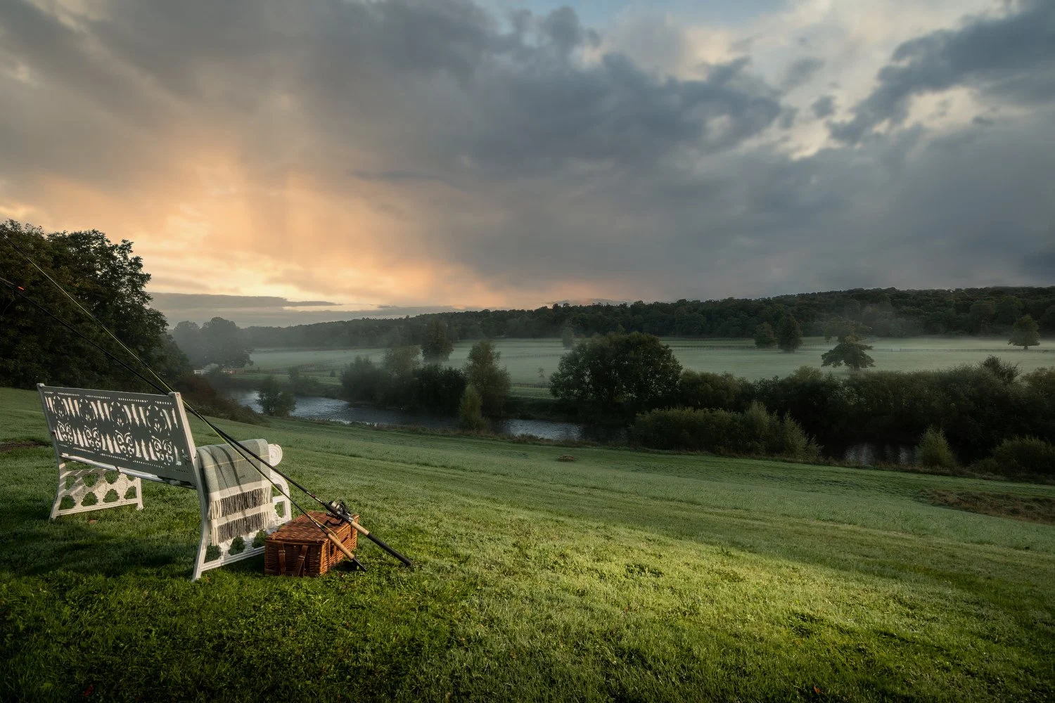 A peaceful outdoor scene at dawn with a bench, fishing rods, and a picnic basket on a grassy hill overlooking a river, with trees, fog, and a cloudy sky.