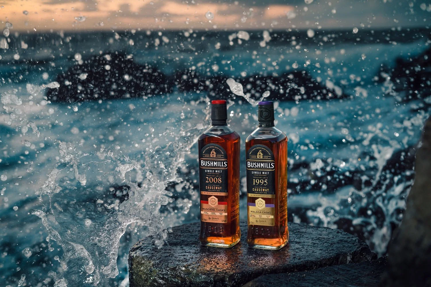 Two bottles of Bushmills single malt whiskey placed on a rock beside water with splashing waves.