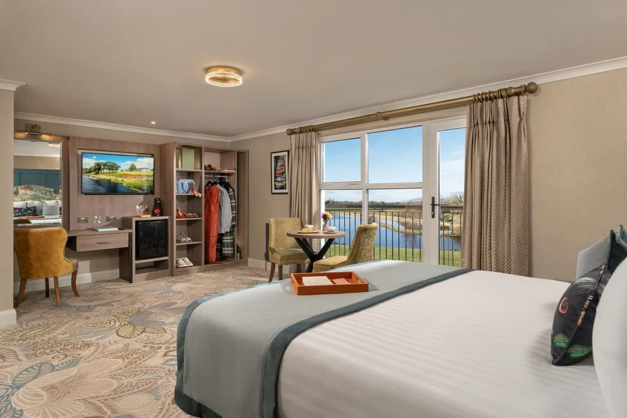 A hotel room with a bed, a small dining table with two chairs, a wardrobe, a flat-screen TV, and a view of a pond or lake outside through large windows.