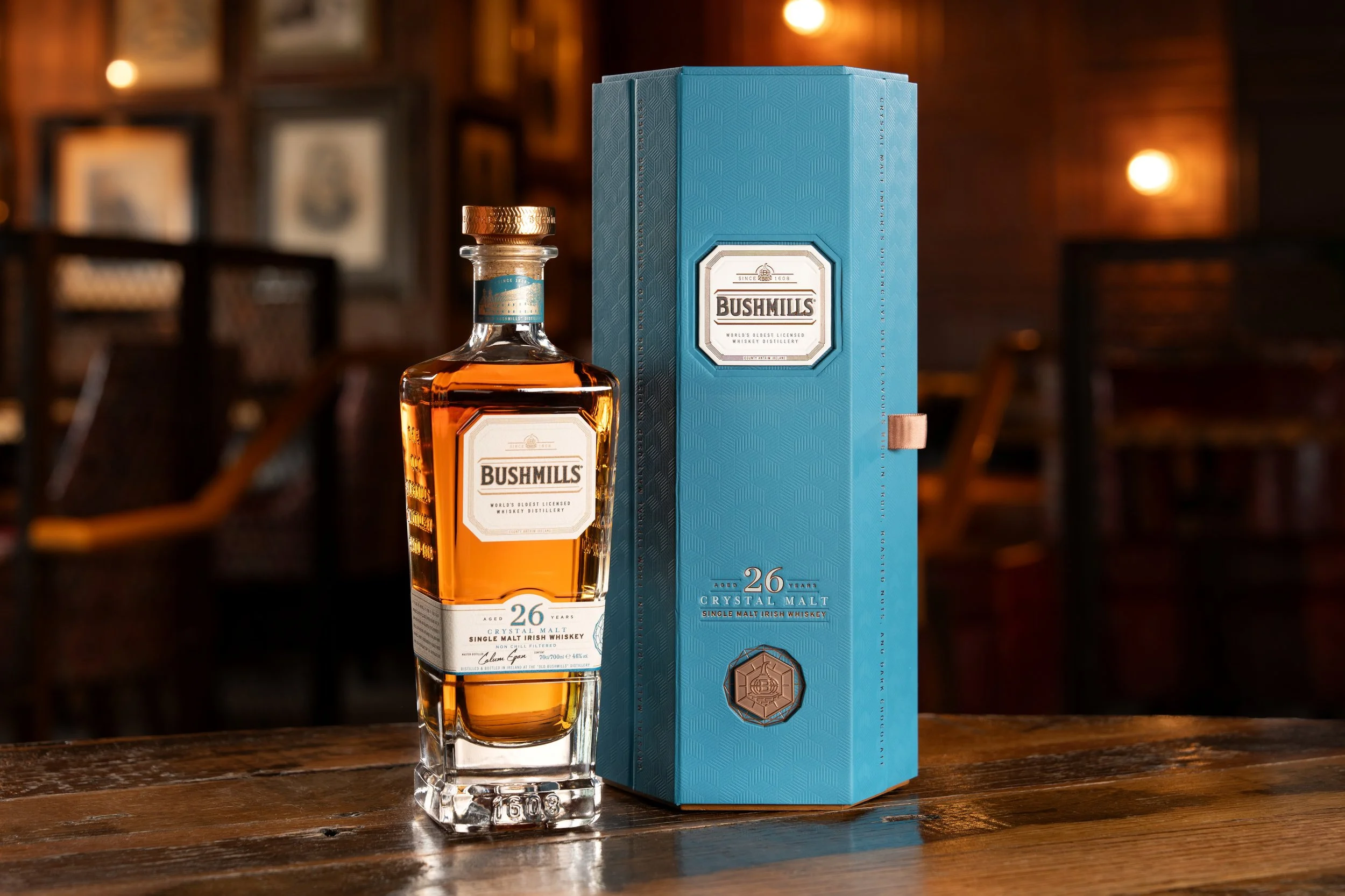 A bottle of Bushmills 26-year-old single malt Irish whiskey next to its blue packaging box on a wooden table in a dimly lit bar.