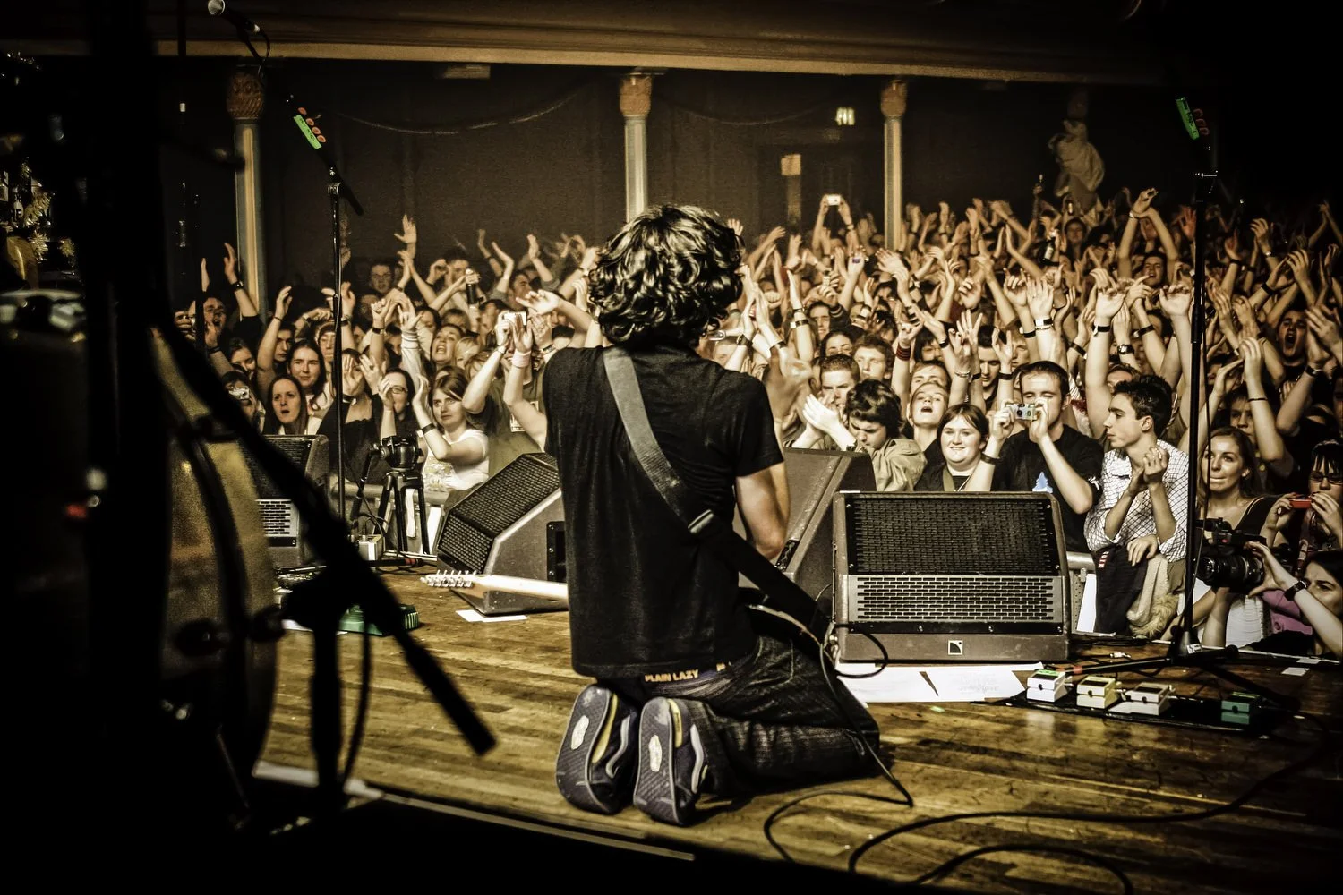 Musician kneeling on stage with a guitar facing a large, cheering crowd at a concert.