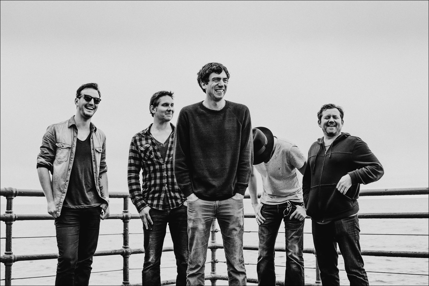 Black and white photo of five men standing on a pier near the water, smiling and laughing. One man is wearing sunglasses, another has a checkered shirt, and a third man is wearing a hoodie. The man in the center is taller and dressed in a dark sweate