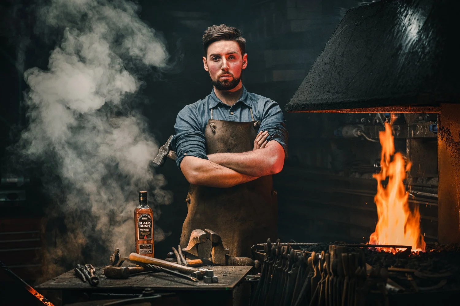A blacksmith in a forge with glowing fire, tools on a workbench, a bottle of Black Bush whiskey, and smoke in the background.
