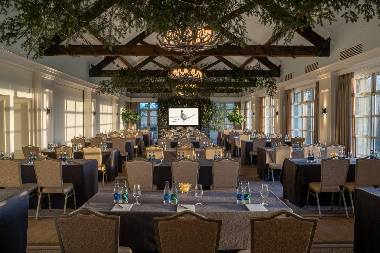 An elegant conference or banquet room with multiple tables covered in dark tablecloths, set with bottled water, glasses, and notepads. The room is well-lit by natural sunlight coming through large windows, with curtains pulled to the sides. The room 