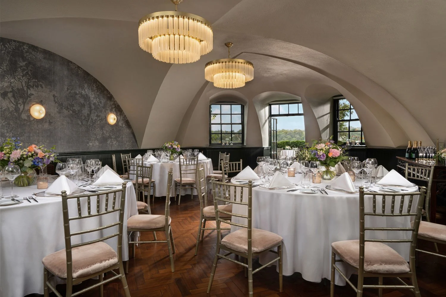Elegant banquet hall with white-draped tables, floral centerpieces, gold chairs, and large windows in an arched ceiling room with modern chandeliers.