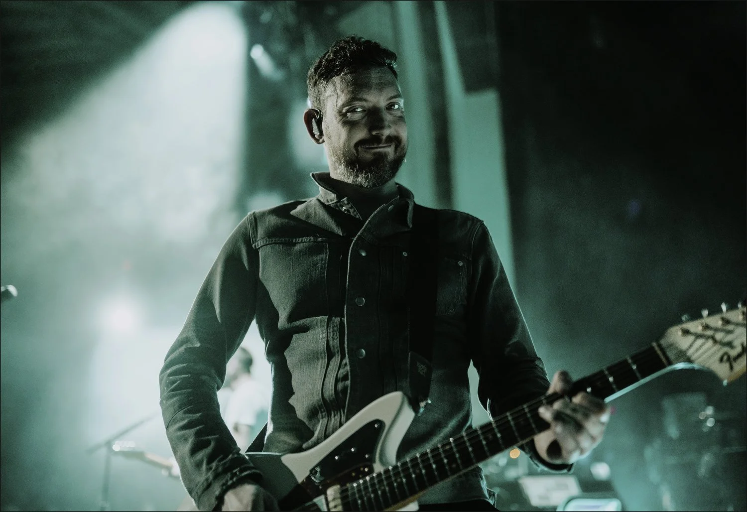 A man playing an electric guitar on stage, wearing a black denim jacket, with studio lights creating a dramatic lighting effect.