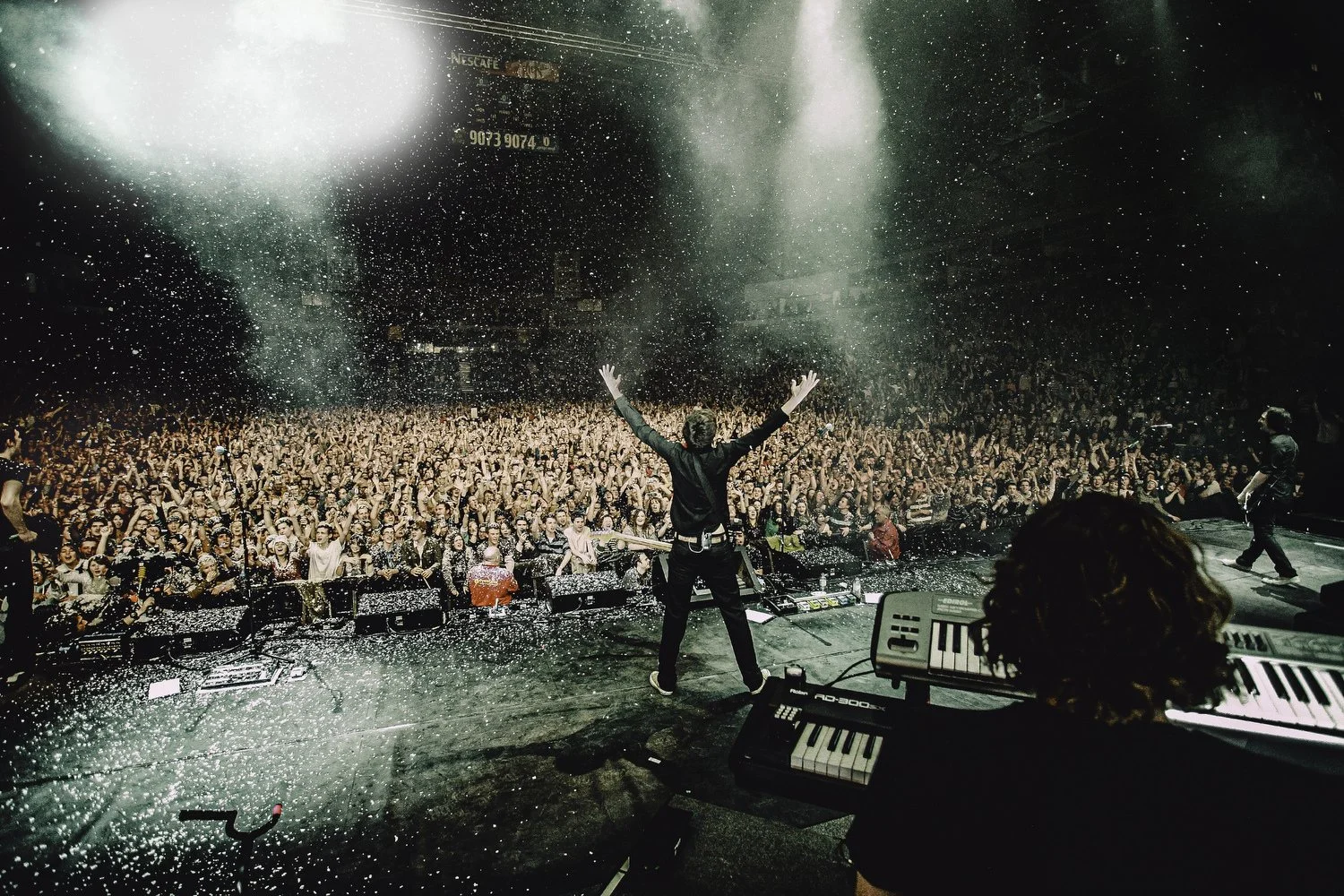 Performer on stage with arms raised, crowd cheering, confetti falling during concert, keyboard and other instruments visible on stage.