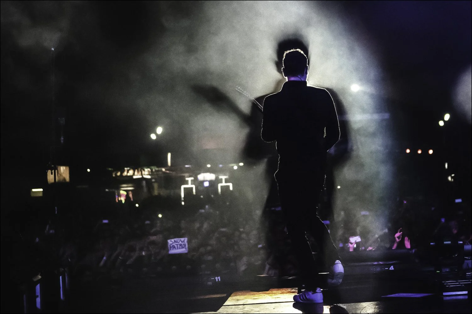 Silhouette of a male performer on stage in front of a large crowd at night, illuminated by stage lights and fog.
