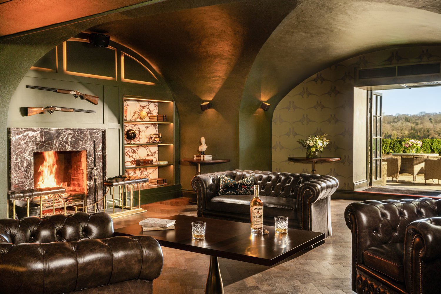 Elegant living room with dark leather sofas, a fireplace with a marble surround, bookshelves with decorative items, a table with a bottle of Jameson whiskey and glasses, and a door opening to an outdoor patio with chairs and greenery.