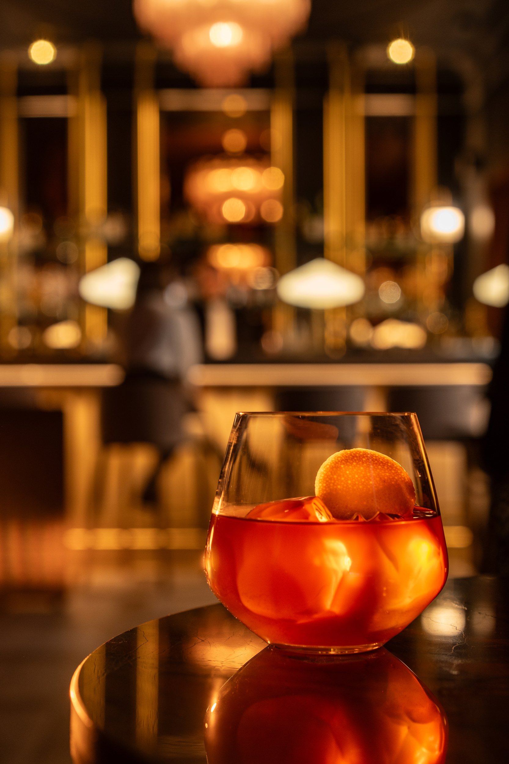 A glass of an orange-colored cocktail with ice and a lemon slice, on a reflective surface in a dimly lit bar or restaurant with blurred background lighting and patrons.