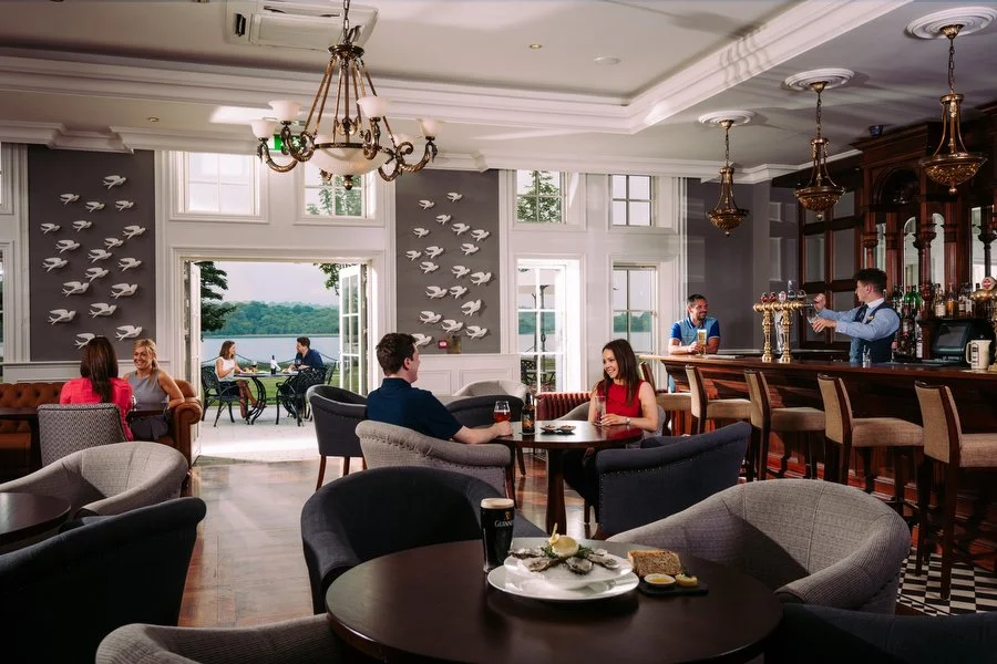 A spacious lounge or bar with high ceilings, white walls, and large windows. People are sitting and chatting at tables, with a view of a lake or water scene outside. The bar area has wooden counters and shelves with bottles, and three bartenders are 