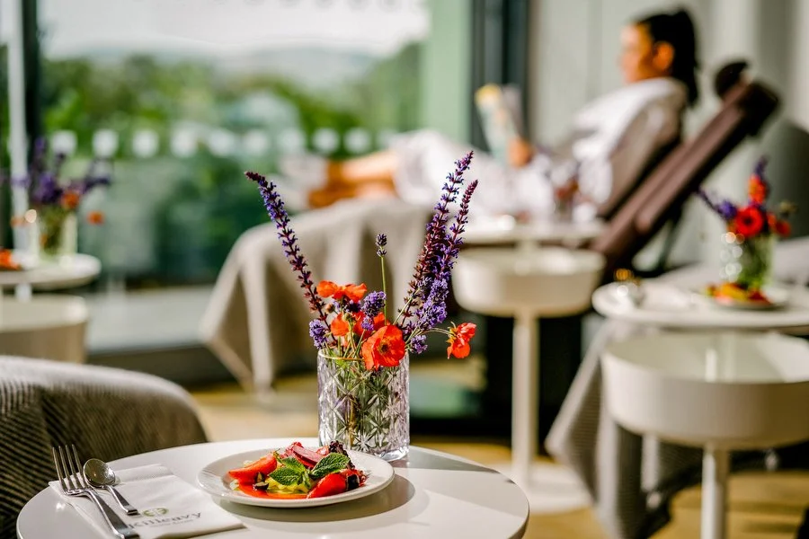 A table with a floral arrangement in a glass vase and a plate of strawberries and mint leaves, set in a bright dining area with large windows. In the background, a woman is relaxing in a chair and reading a book.