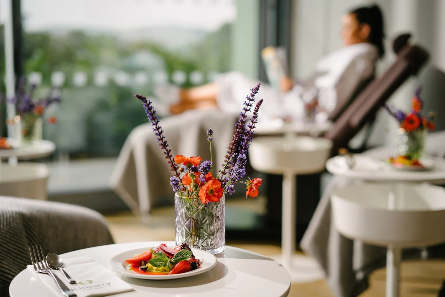 A dining table with a white tablecloth, featuring a plate of salad, a fork, a spoon, a napkin, and a vase of purple and orange flowers, with a woman in a sunlit room in the background.