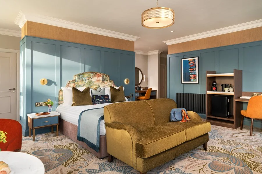 Hotel bedroom with blue and beige decor, bed with floral headboard, green sofa, and wooden furniture.