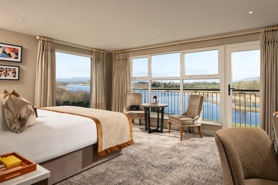 A hotel room with a large window showing a view of a river, trees, and distant hills; a bed with pillows, curtains, and furniture.