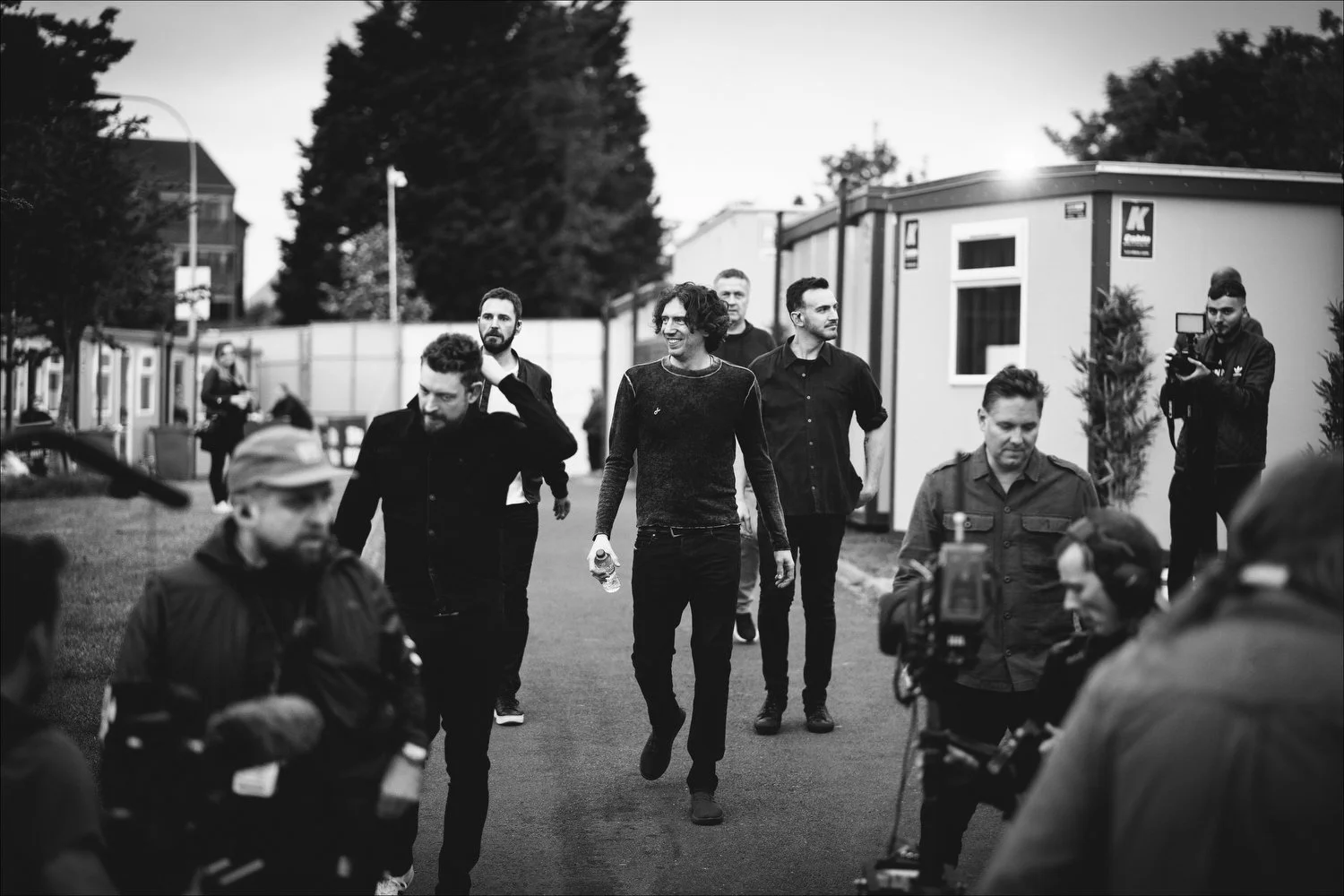 A group of men walking outdoors, some looking at the camera, with a film crew and equipment capturing the scene. Black and white photo.