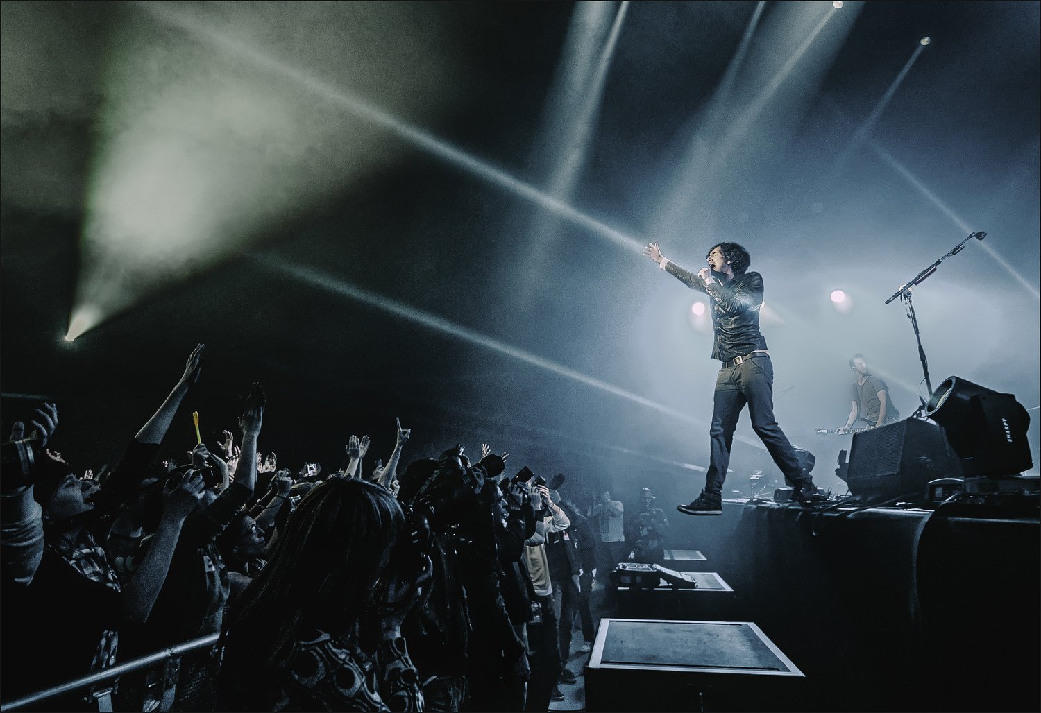 A singer on stage reaching out towards a crowd during a concert, with a keyboardist in the background, stage lights illuminating the scene, and the audience with hands raised.