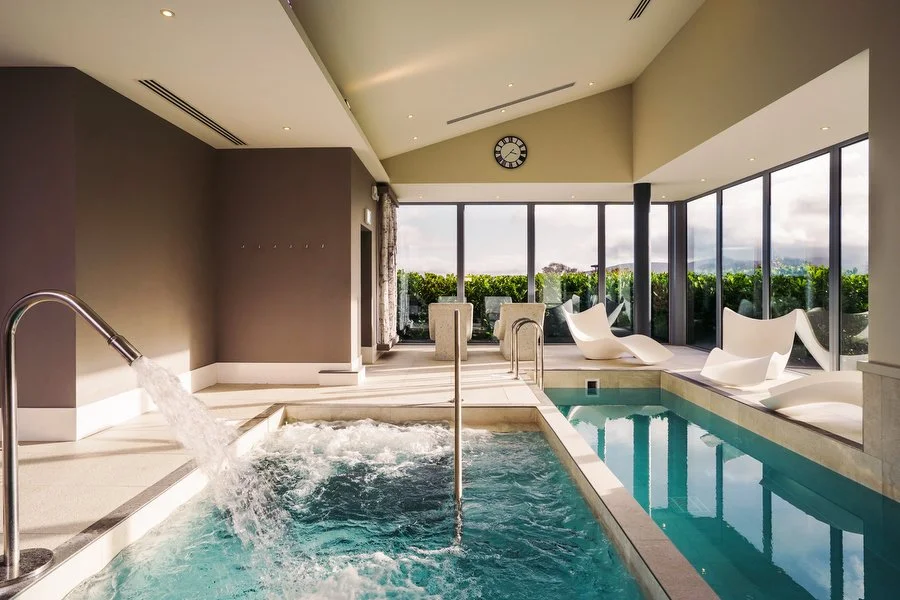 Indoor spa area with a pool, white lounge chairs, and floor-to-ceiling windows showing an outdoor view, with a clock on the wall Killeavy Estate