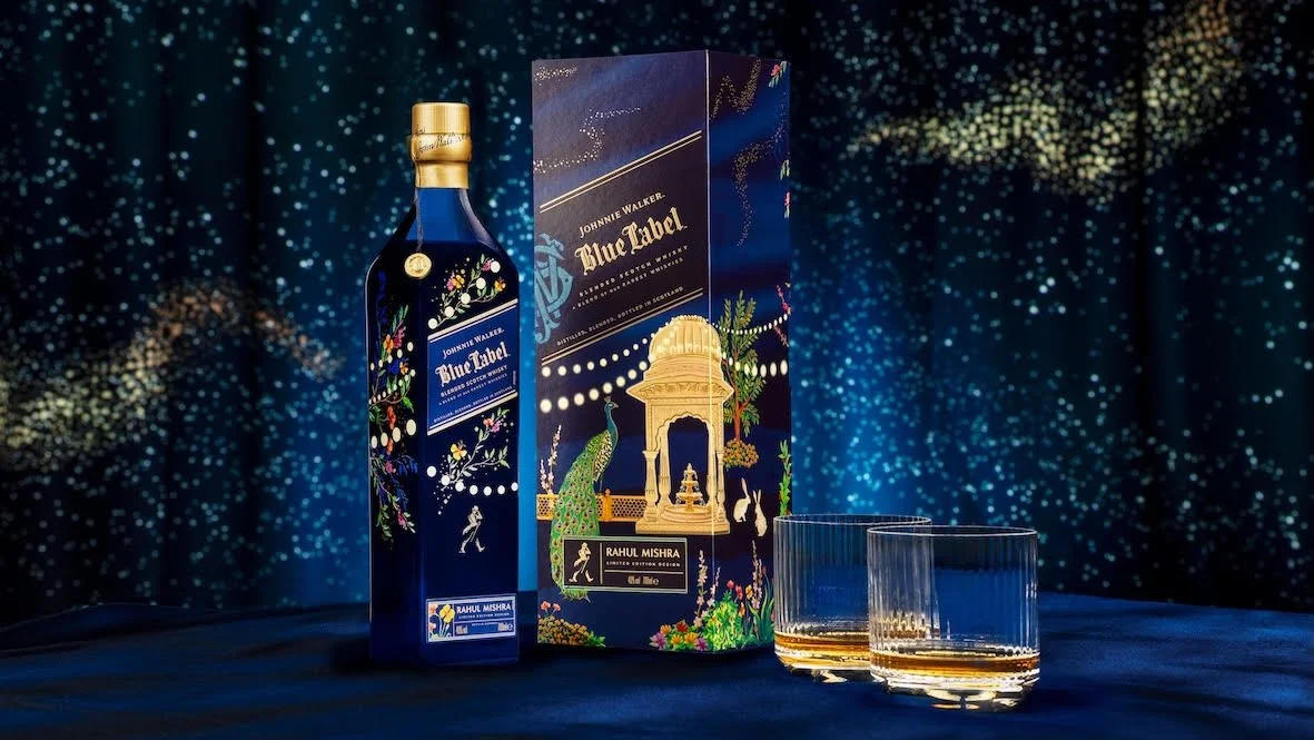 A bottle of Johnnie Walker Blue Label whisky, its packaging box, and two glasses of whisky on a dark surface, with a starry, galaxy-like background.