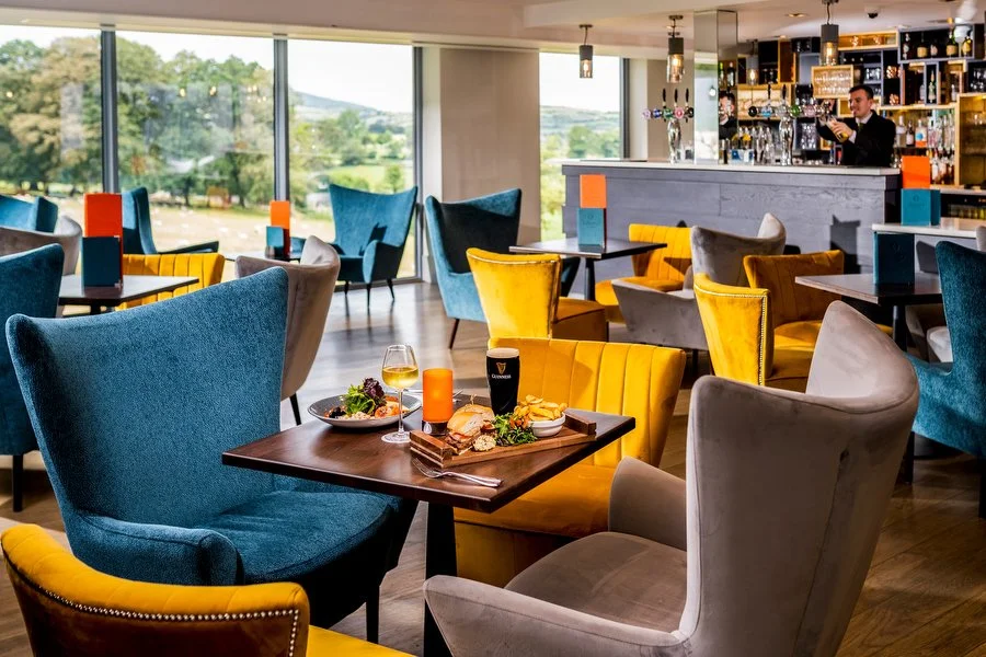 A modern, bright restaurant or lounge area with colorful chairs, a bar in the background, large windows showing a scenic outdoor view, and a table with drinks and food.