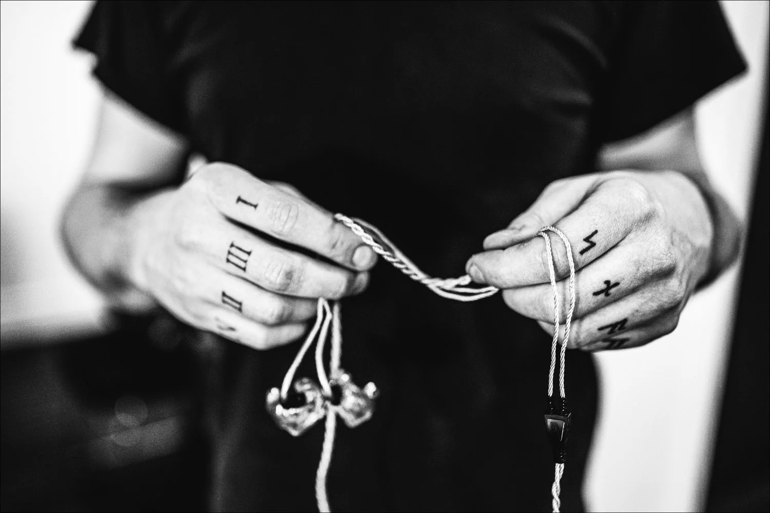 Close-up of tattooed hands holding earphone and a string of jewelry, with tattoos of Roman numerals and letters.