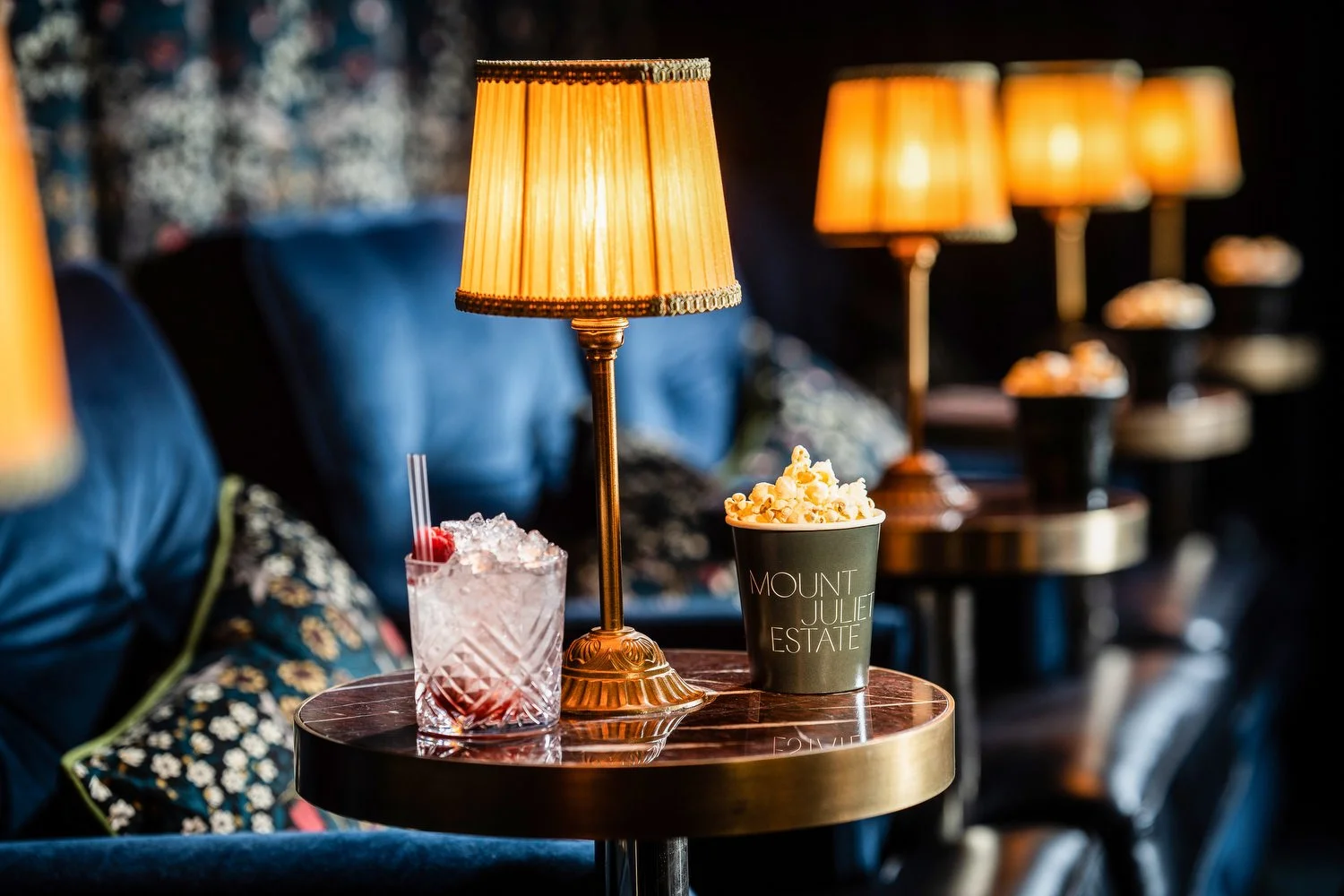 A round table with a table lamp, a glass of cocktail with a cherry and ice, and a bucket of popcorn, in a dimly lit lounge or bar with leather couches and multiple lamps in the background.
