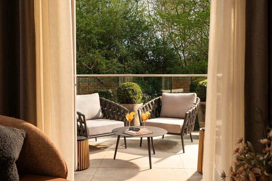 Balcony with two grey cushioned chairs, a small round table with drinks and a book, potted bushes, and a glass railing overlooking trees.